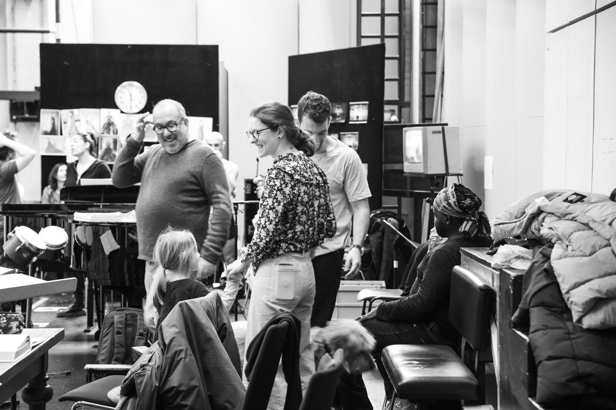 Black-and-white photo from the rehearsal room: Anisha Bondy laughs in conversation with Barrie Kosky, surrounded by cast members and a child.