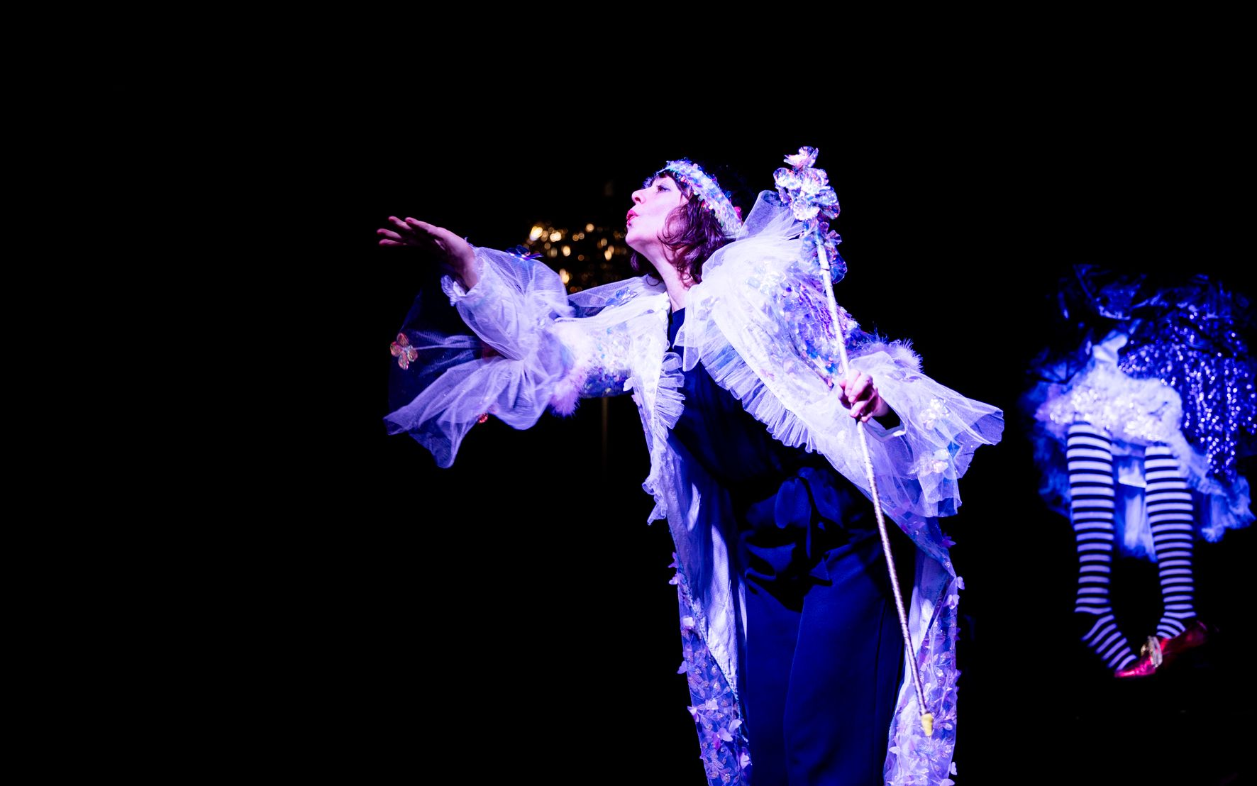 A performer in a white feathered costume spreads her arms, beside her a striped puppet figure.