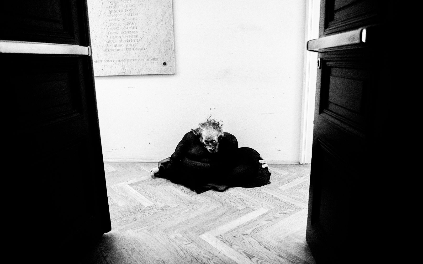 Black-and-white: a costumed figure crouches on the floor between two open doors.