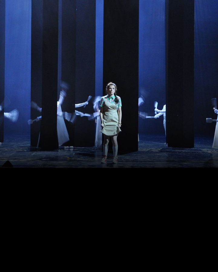 A girl standing among tall dark pillars and ghostly figures with brooms.