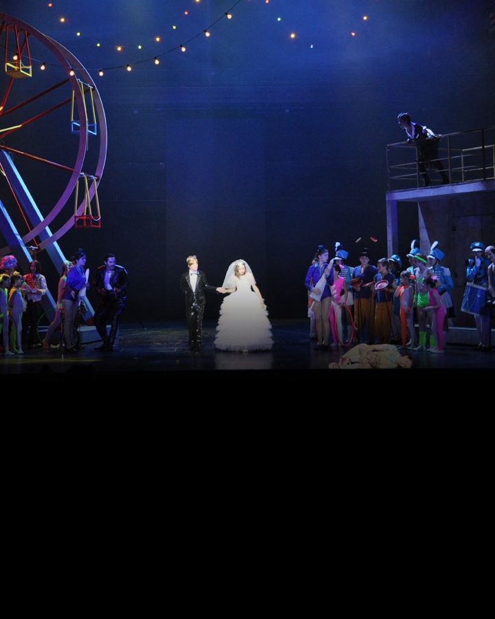 A wedding couple in front of a colourful ferris wheel, surrounded by the ensemble.