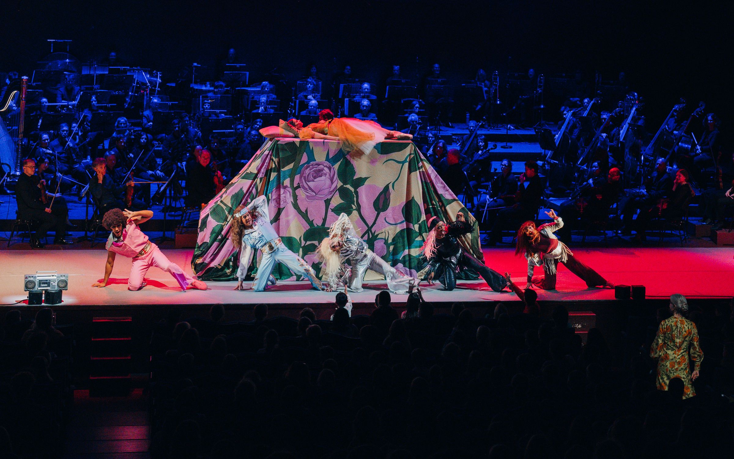 Dancers in front of the rose tent, the orchestra in blue light behind them.