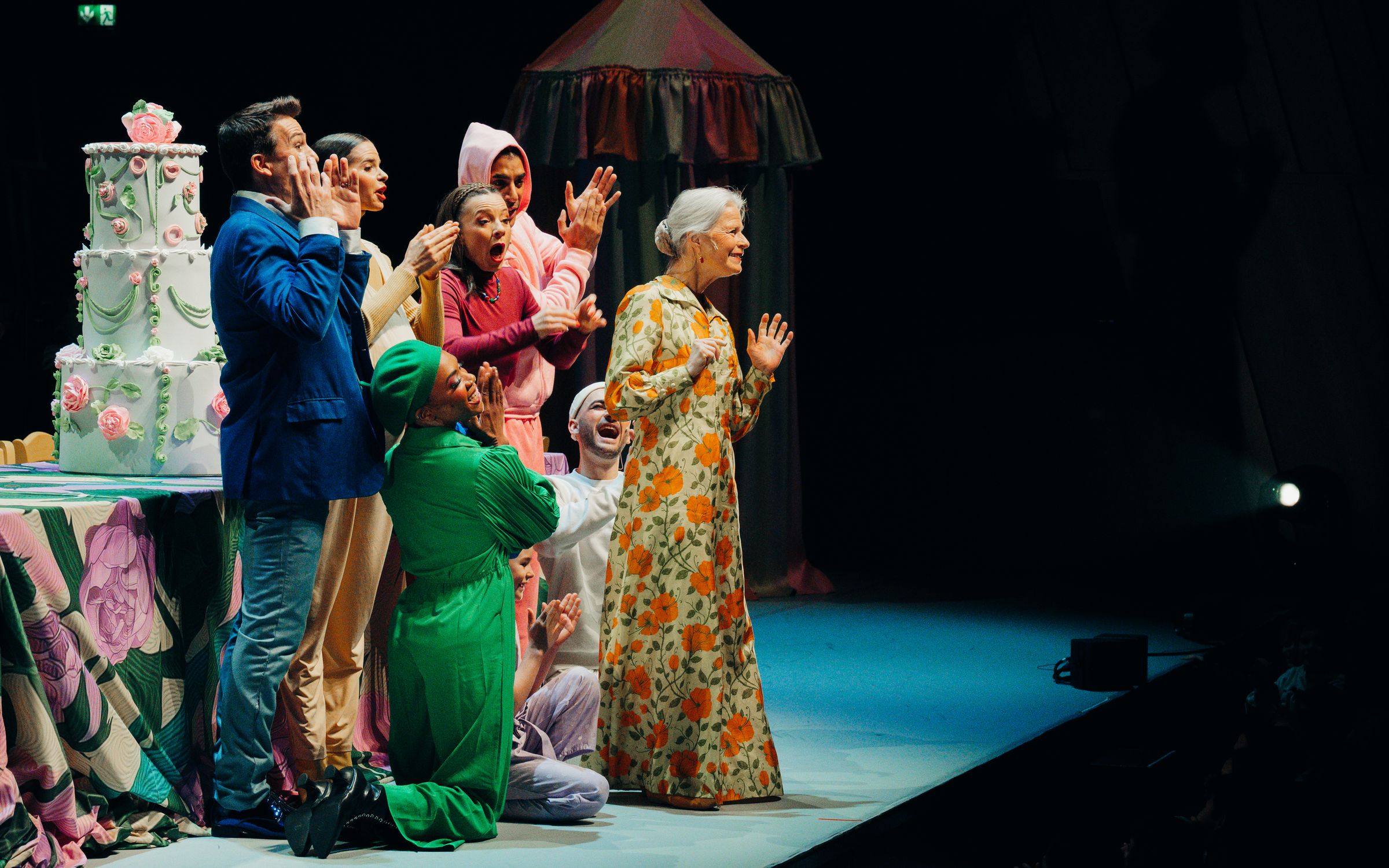 Performers in colourful costumes gather excitedly around an old woman in a floral dress on stage.