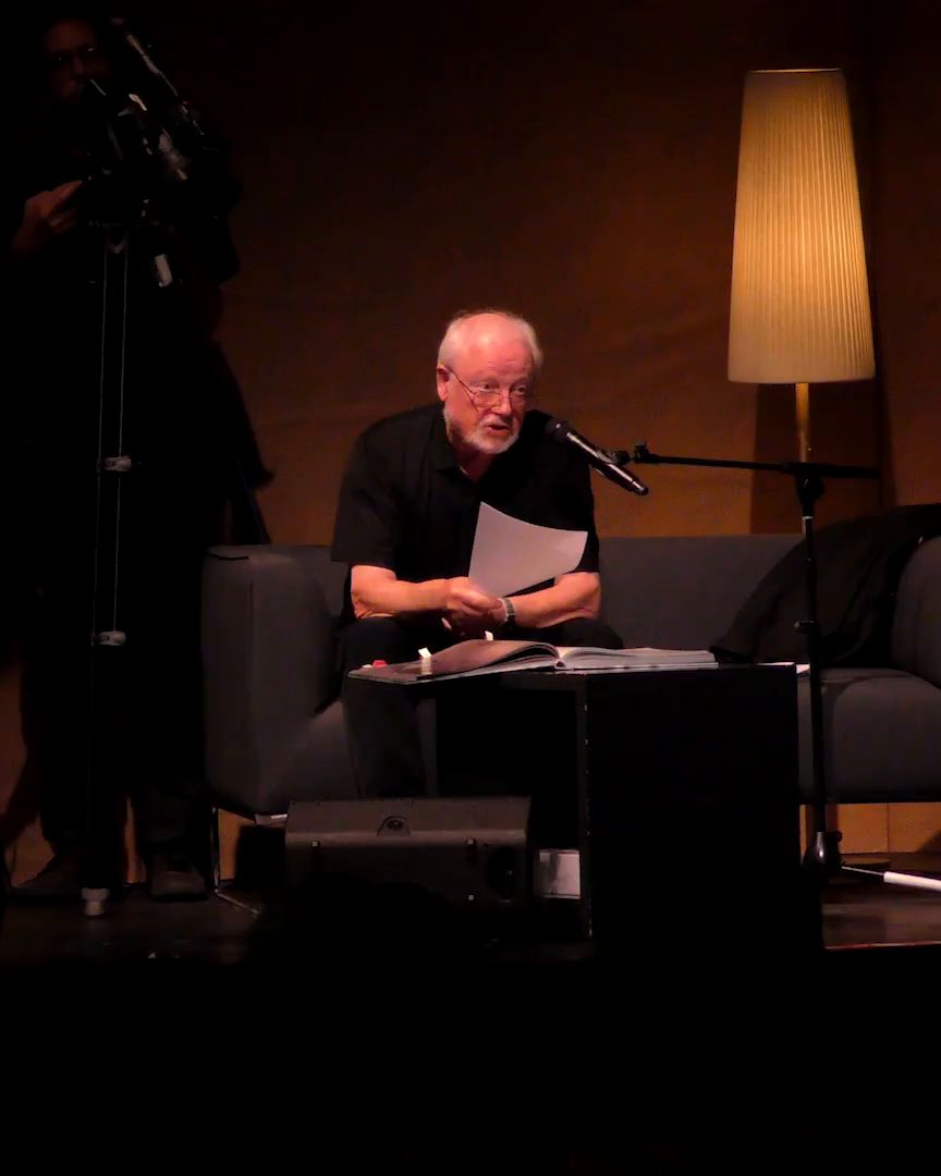A man reads from a sheet of paper, seated under a floor lamp.