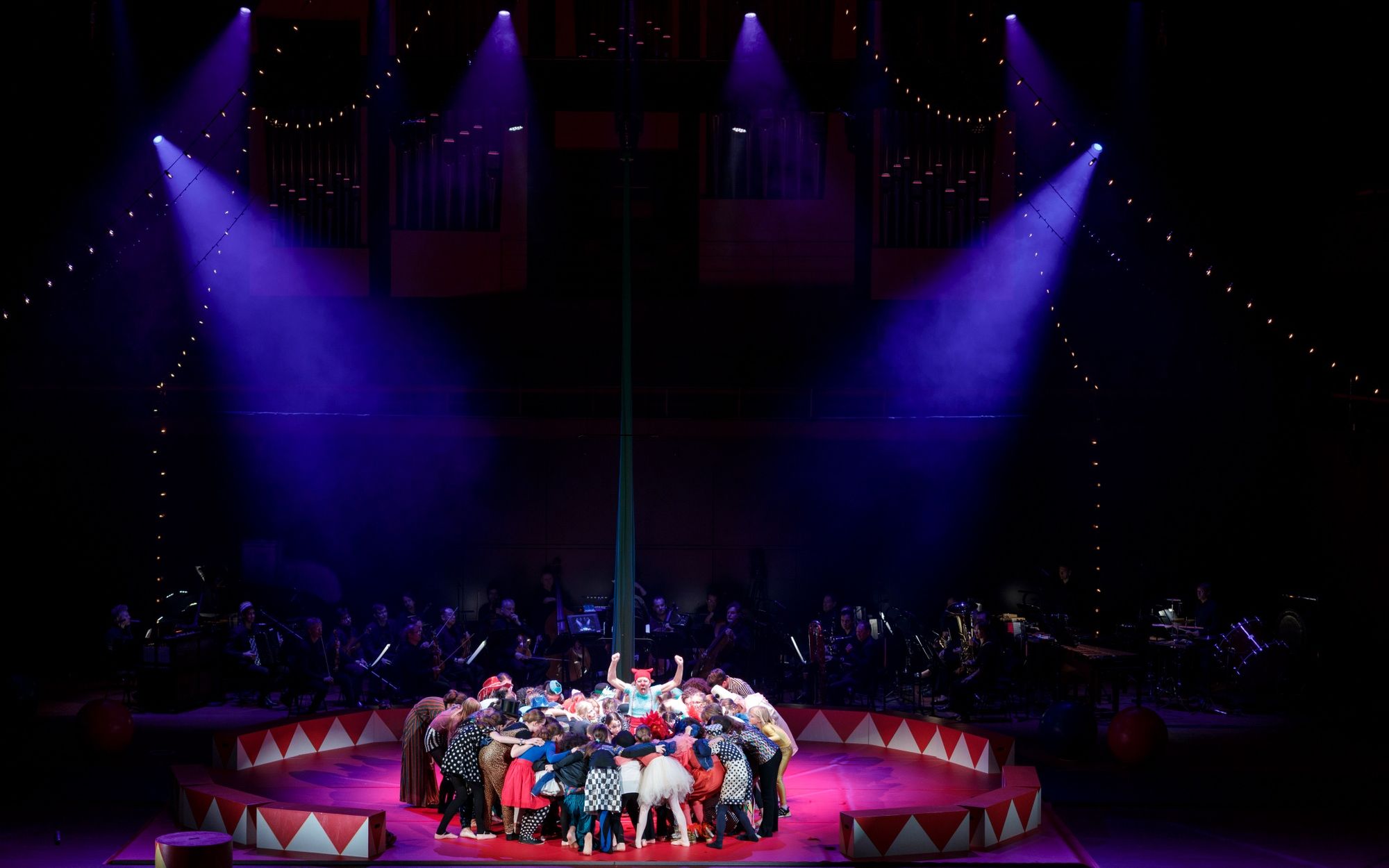 Singers and circus artists gather in the circus ring under purple spotlights.