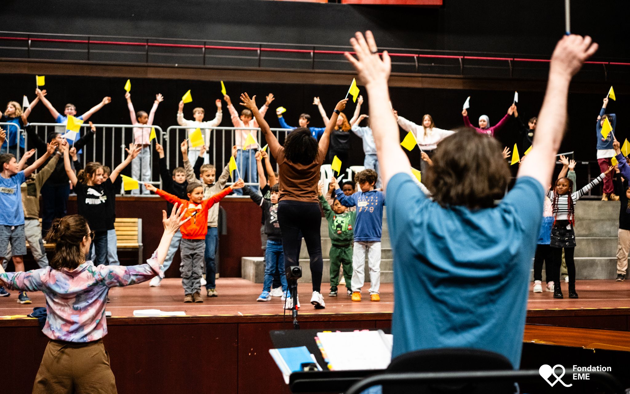 Children on stage throw their arms in the air, waving small yellow flags.