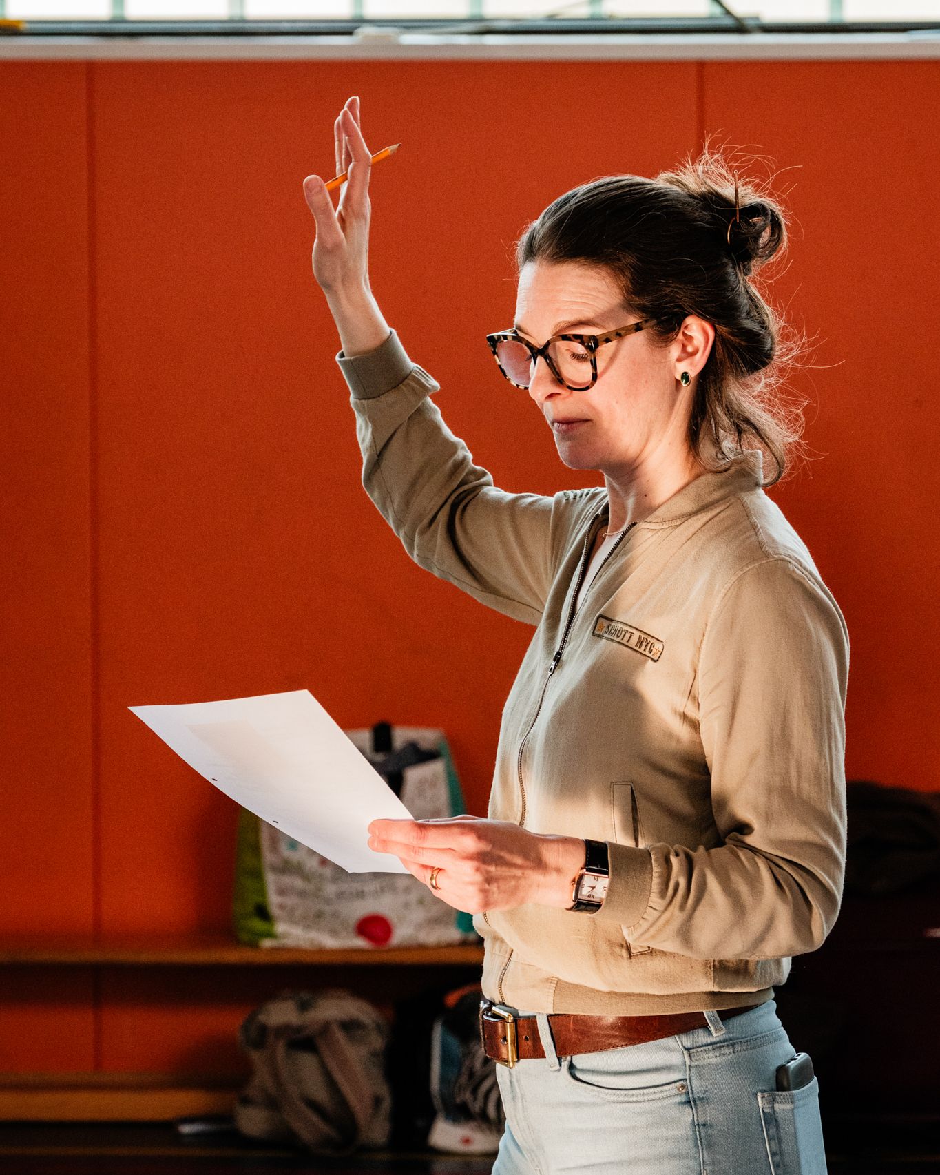 At a rehearsal, a woman raises her arm, a sheet of paper in her hand, in front of an orange wall.