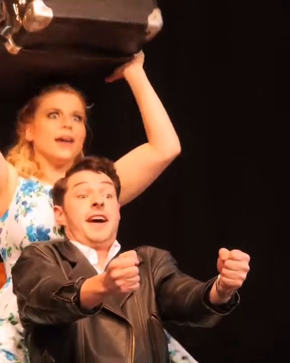 Both singers at the front of the stage, he with clenched fists as if steering.