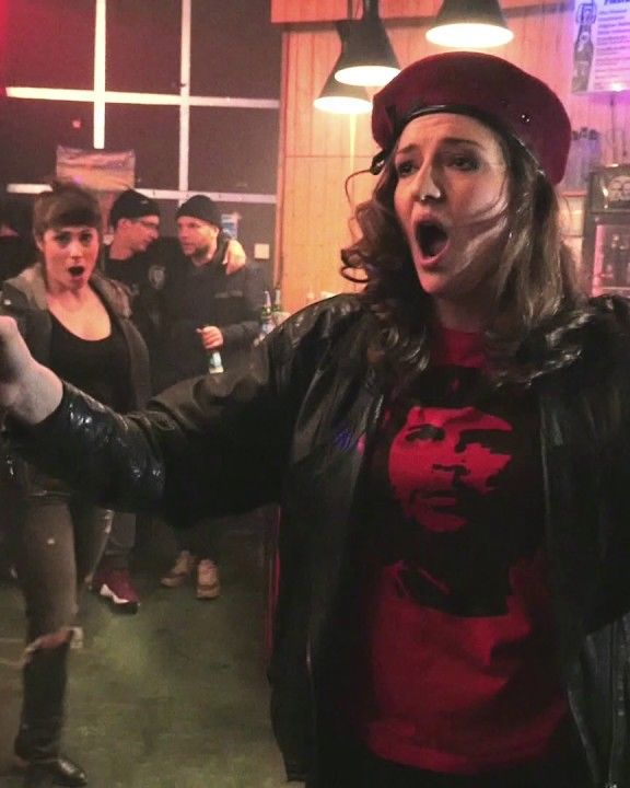A singer in a beret and leather jacket sings with outstretched arms among the audience of a pop-up opera.